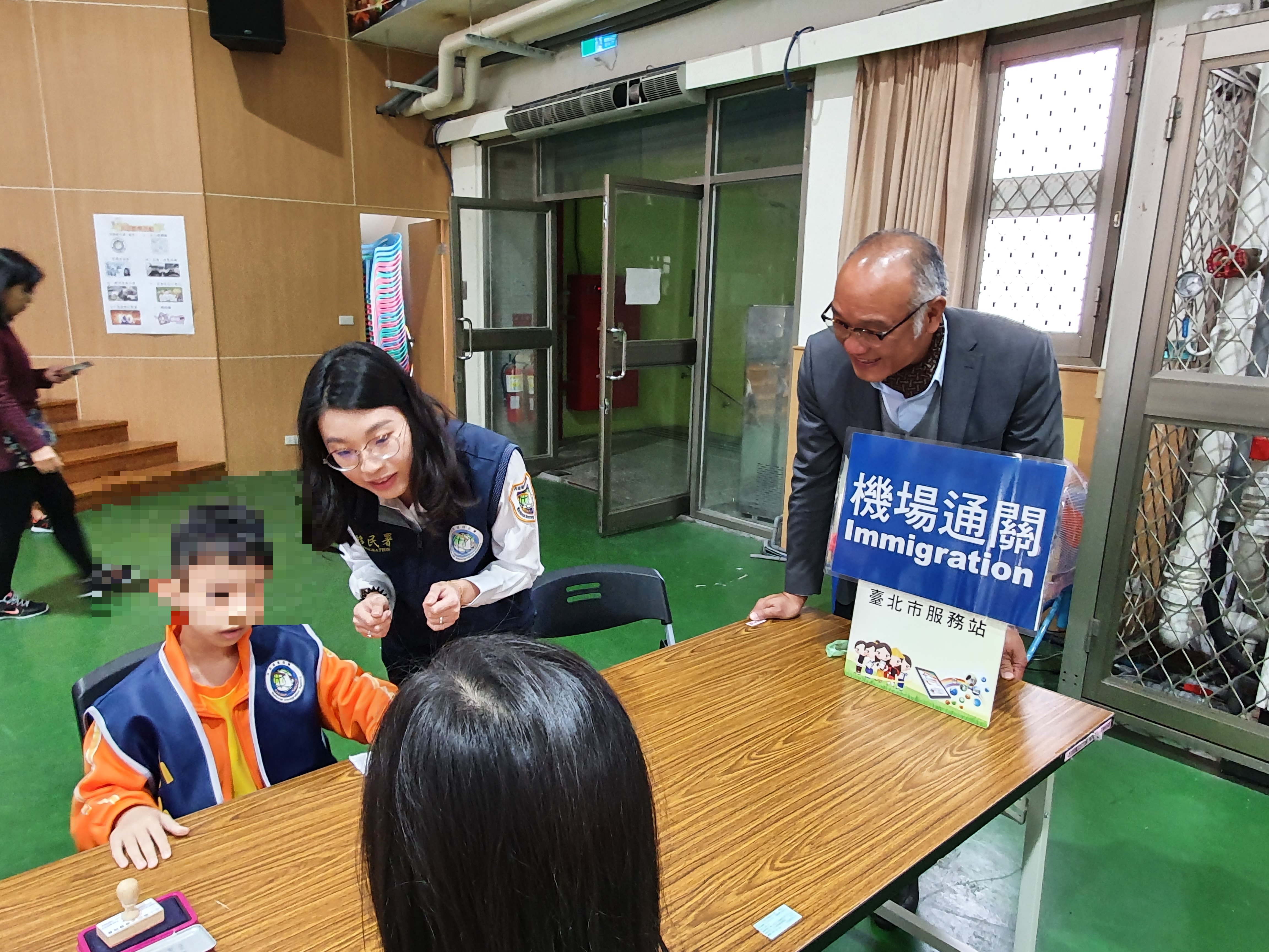 移民官張瓈云（中）教學童機場通關查驗護照。(右為明德國小校長涂振洋) (移民署臺北市服務站提供)